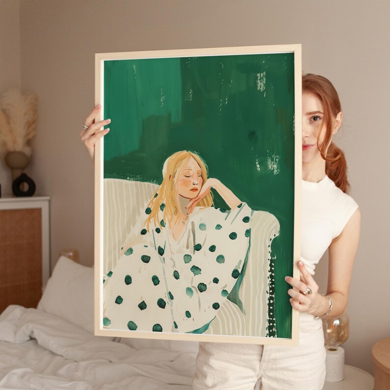 May include: A framed art print featuring a watercolor illustration of a woman with blonde hair, wearing a white robe with large green polka dots, seated on a white sofa against a green background.