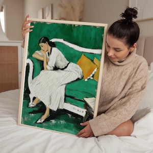 以下が含まれることがあります： 白いドレスを着た女性が黄色のクッションが付いた緑色のソファに座っている、額装されたアートプリント。 アートワークは明るい木製のフレームとヴィンテージの美学を持っています。 女性は本を読んでいます。