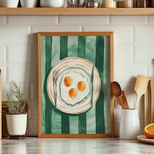 May include: A framed watercolor painting of a plate of eggs with three sunny-side up eggs on a green and white striped background.