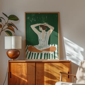 May include: A framed illustration of a person sitting on a striped green and white beach towel. The person is wearing a striped green and white dress and has their hands on their head. The background is a solid green color.