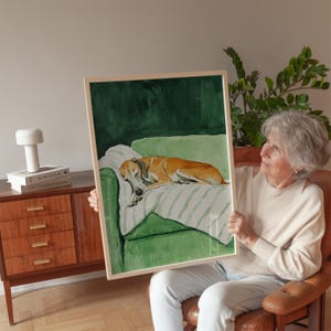 Puede incluir: Una pintura al agua enmarcada de un perro durmiendo en un sofá verde. El sofá tiene un cojín a rayas blanco y verde. La pintura la sostiene una persona con un suéter blanco.