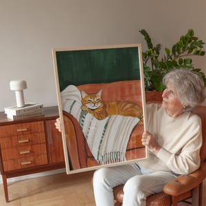 Könnte beinhalten: Ein gerahmtes Gemälde eines roten Katers, der auf einem Sofa mit grünem Hintergrund entspannt. Die Katze schaut den Betrachter mit einem entspannten Ausdruck an.