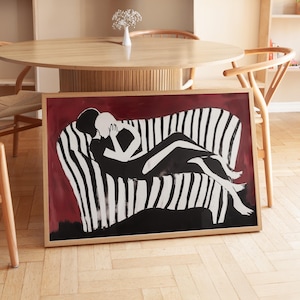 May include: A framed art print depicting a black and white illustration of two figures on a striped sofa. The artwork has a red background and is in a natural wood frame. The print is displayed in a room with wooden furniture.