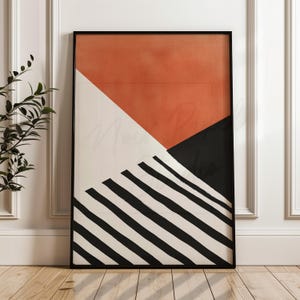 May include: Abstract art print in a black frame. The artwork features geometric shapes: a burnt orange triangle, a white triangle, and a black triangle. Diagonal black and white stripes complete the design. The print is displayed on a wooden floor.