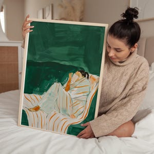 以下が含まれることがあります： ストライプのソファでくつろぐ人の抽象的なイラストが描かれた、額入りのアートプリント。アートワークは緑色の背景で、人物はストライプの毛布に覆われています。フレームは明るい木の色です。