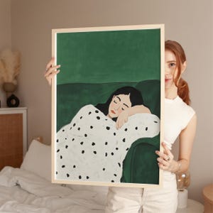 May include: A framed art print featuring a woman resting on a green sofa, draped in a white blanket with black polka dots. The artwork has a green background and is held by a person. The frame is a light wood color.