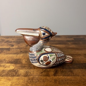 May include: A ceramic pelican figurine featuring a brown beak and a cream-coloured body decorated with intricate hand-painted patterns in blue, green, and brown. The design includes floral motifs, dots, and geometric shapes across the wings and neck.