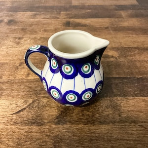 May include: A small, round creamer jug with a handle and spout. The ceramic jug is white with a blue band featuring a repeating pattern of blue circles with green and red centres. The handle is blue with a matching pattern.
