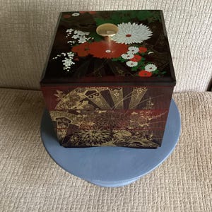 Peut inclure: Une boîte décorative à plusieurs niveaux avec un motif floral. La boîte est principalement rouge foncé avec des accents dorés et un couvercle avec des fleurs blanches, rouges et vertes. Elle a un bouton doré et repose sur un support bleu clair en forme de cœur.