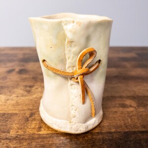 May include: A handmade ceramic vase with a cream-coloured glaze and a decorative leather tie. The vase has a unique shape and texture, with a detailed rim and a leather accent. The vase is a decorative item.