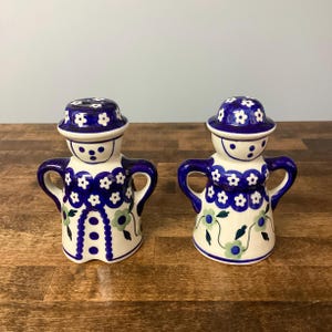 May include: Two ceramic figurines with a white base, blue floral patterns and accents. Each has a hat-like top and arms, with a face design. The design is in a folk art style. The height of each figurine is approximately 10 cm.