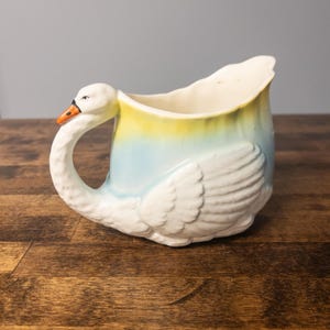 Peut inclure: Un pichet ou pot à lait en céramique en forme de cygne, doté d'un corps blanc avec des détails d'ailes moulés, une anse en forme de cou courbé et un dégradé passant du bleu clair au jaune sur le bord supérieur, avec un bec orange et un œil noir.