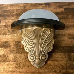 May include: An Art Deco style wall sconce. It features a brass shell-shaped base, a black rim, and a light blue, dome-shaped glass shade. The sconce is designed to be mounted on a wall.