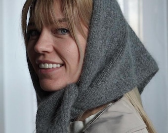 May include: A gray knitted head covering, worn over the head and tied under the chin. The head covering is made of a soft, textured material. The person is wearing a light-colored coat.