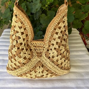 May include: A hobo-style shoulder bag crafted from crocheted yarn in alternating shades of beige and tan. The bag features an open-work granny square pattern, a solid tan interior lining, and a tan strap attached with silver-toned metal hardware.