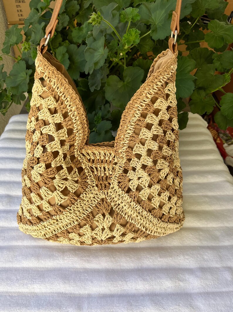 May include: A hobo-style shoulder bag crafted from crocheted yarn in alternating shades of beige and tan. The bag features an open-work granny square pattern, a solid tan interior lining, and a tan strap attached with silver-toned metal hardware.