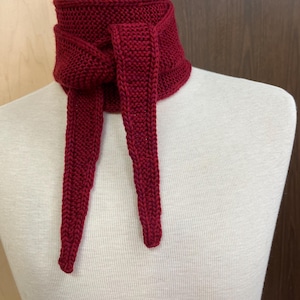 May include: A knitted scarf in a deep red hue, displayed on a mannequin. The scarf features a thick, textured knit with long, dangling ends. The design wraps around the neck and ties in the front, offering a warm and stylish accessory.