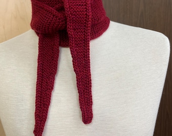 May include: A knitted scarf in a deep red hue, displayed on a mannequin. The scarf features a thick, textured knit with long, dangling ends. The design wraps around the neck and ties in the front, offering a warm and stylish accessory.