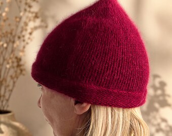 May include: A knitted burgundy hat with a pointed top. The hat is made of a soft, textured yarn and is worn by a person with blonde hair. The hat is a unique and stylish accessory.