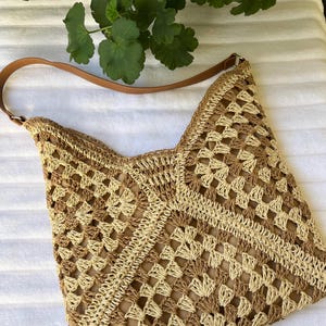 May include: A crochet shoulder bag featuring a geometric granny square pattern in alternating shades of beige and tan. The bag includes a smooth brown leather-like strap attached with silver-toned metal hardware and a solid tan interior lining.