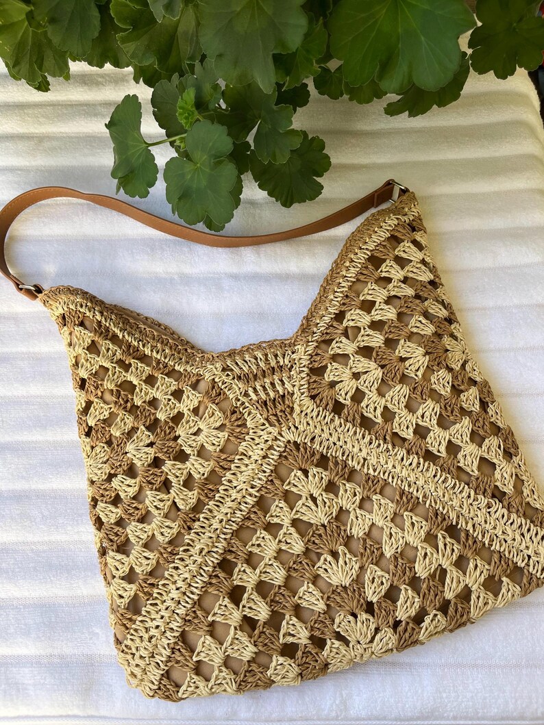 May include: A crochet shoulder bag featuring a geometric granny square pattern in alternating shades of beige and tan. The bag includes a smooth brown leather-like strap attached with silver-toned metal hardware and a solid tan interior lining.