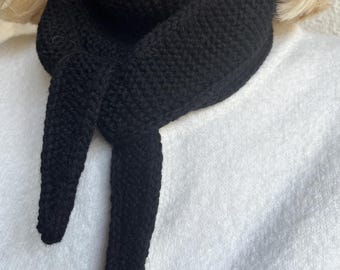 May include: A black knitted scarf with a textured appearance. The scarf is wrapped around the neck and has two long, pointed ends. The scarf is made of a thick yarn and is a solid black color. The scarf is worn over a white garment.