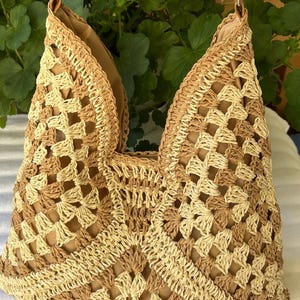 May include: A crochet handbag featuring a geometric pattern in alternating shades of beige and tan. The bag has a structured open top, a solid tan interior lining, and a thin tan strap attached with silver-toned metal hardware rings.