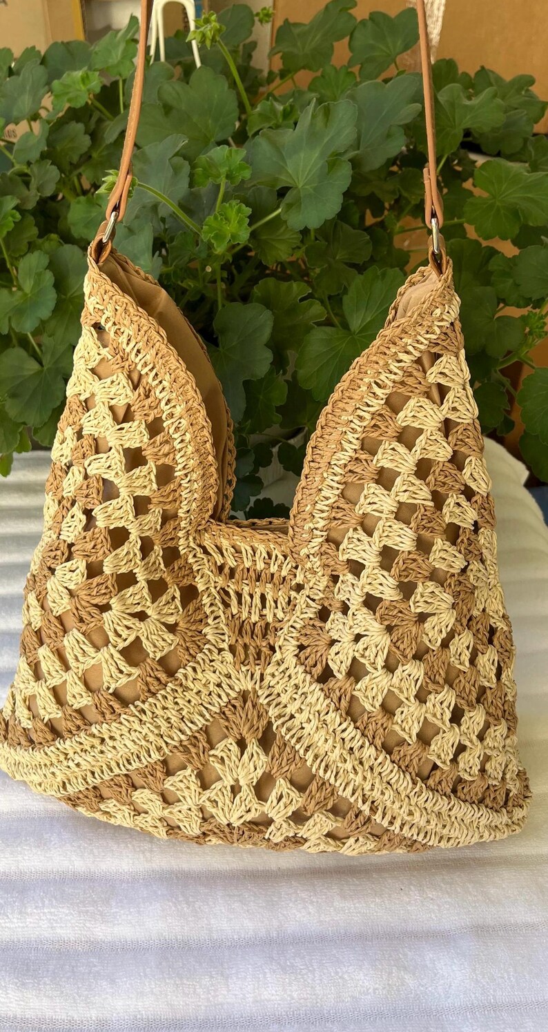 May include: A crochet handbag featuring a geometric pattern in alternating shades of beige and tan. The bag has a structured open top, a solid tan interior lining, and a thin tan strap attached with silver-toned metal hardware rings.