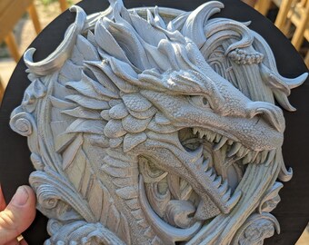 Puede incluir: Escultura detallada de un dragón gris montada sobre una placa de madera negra y circular. El dragón está intrincadamente tallado con escamas, dientes afilados y detalles fluidos. La obra de arte es una escultura en relieve, creando un efecto 3D.