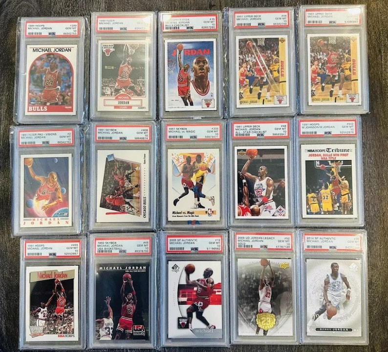 May include: A collection of 12 Michael Jordan basketball trading cards in protective plastic cases. The cards feature various images of Jordan playing basketball, including a card with the text "NBA Hoops Tribune Jordan Bulls Win First Title".