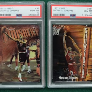 May include: Two 1997 Finest Michael Jordan basketball cards, both graded Gem Mint 10 by PSA. The card on the left features Jordan in a red and black Bulls uniform, with the word "Finishers" in gold lettering. The card on the right features Jordan in a red and black Bulls uniform, with the word "Showstoppers" in gold lettering.