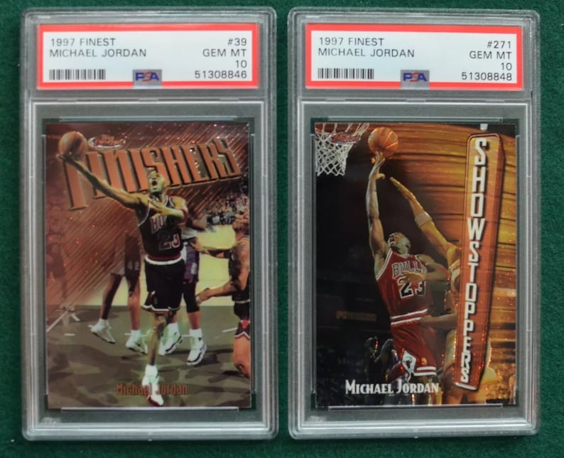 May include: Two 1997 Finest Michael Jordan basketball cards, both graded Gem Mint 10 by PSA. The card on the left features Jordan in a red and black Bulls uniform, with the word "Finishers" in gold lettering. The card on the right features Jordan in a red and black Bulls uniform, with the word "Showstoppers" in gold lettering.