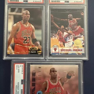 May include: Three graded trading cards of Michael Jordan, a retired American professional basketball player. The cards are from the 1992 Stadium Club, 1993 Hoops, and 1994 Emotion sets. Each card features a different image of Jordan in his Chicago Bulls uniform. The cards are graded GEM MT 10 by PSA.