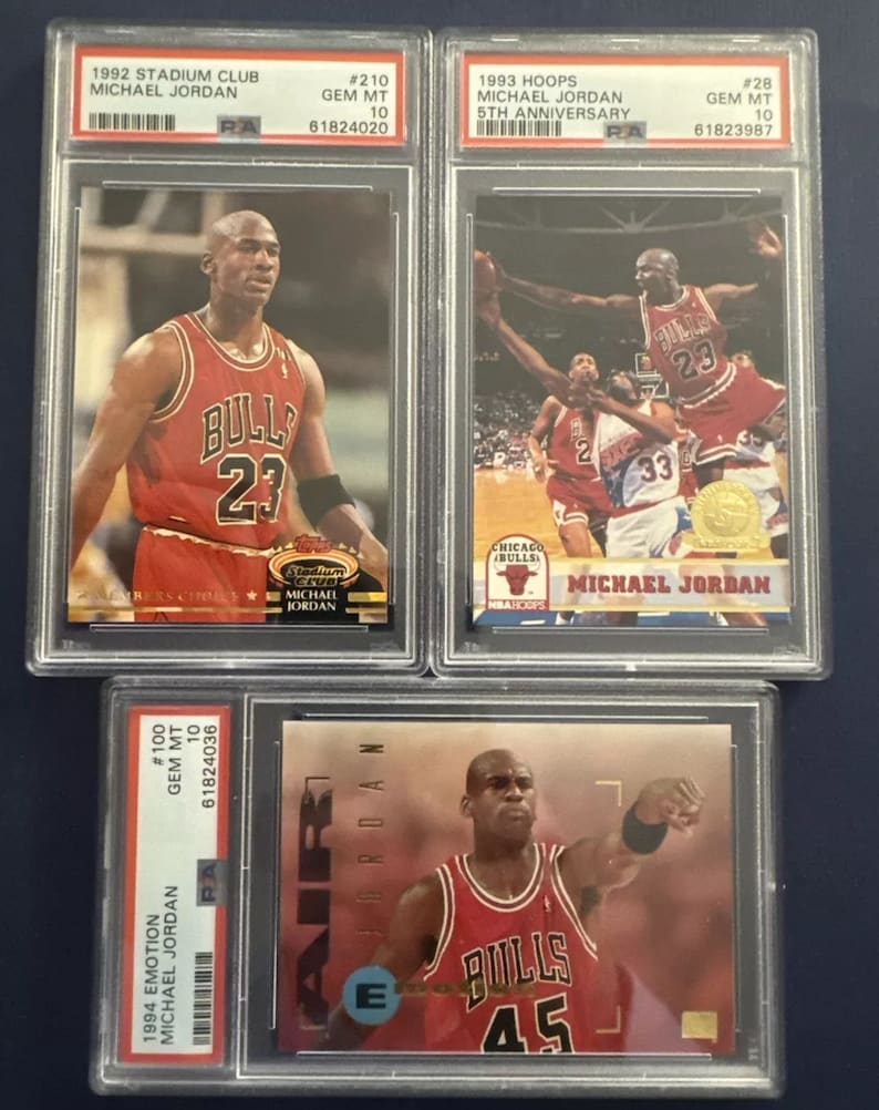 May include: Three graded trading cards of Michael Jordan, a retired American professional basketball player. The cards are from the 1992 Stadium Club, 1993 Hoops, and 1994 Emotion sets. Each card features a different image of Jordan in his Chicago Bulls uniform. The cards are graded GEM MT 10 by PSA.