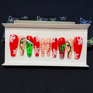 May include: Set of ten red and white Christmas-themed press-on nails. The nails feature a Grinch design, candy canes, snowflakes, and Christmas lights.