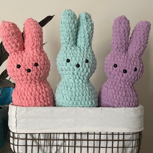 Puede incluir: Tres peluches de conejo de crochet en una cesta de alambre. Los conejos son rosa, azul claro y morado.