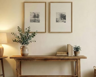 May include: A long wooden console table holds a ceramic vase with eucalyptus branches, a stack of books, and a small potted plant. Two framed black and white landscape prints hang on the wall above the table, next to a table lamp.