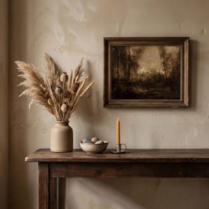 May include: A dark wooden table holds a matte beige vase filled with dried pampas grass, wheat, and poppy pods. Beside it sits a small ceramic bowl containing smooth stones and a yellow taper candle in a metal holder, beneath a framed landscape painting.