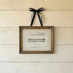 May include: A framed black and white seascape print with a gold beaded frame, suspended by a black satin ribbon. The print shows a coastal scene with sailboats and waves. The frame is rectangular.
