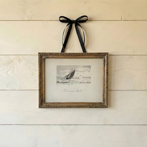 May include: A framed nautical print of a sailboat at sea, with a gold-toned frame and a black ribbon for hanging. The black and white print has the words "Weiland Harbor" at the bottom, and is a classic maritime image.