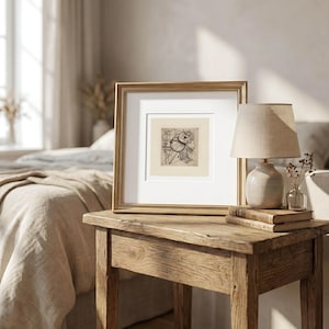 May include: A framed abstract art print in a gold-colored frame. The artwork is a black and white sketch. The frame sits on a rustic wooden side table, next to a small lamp with a beige lampshade and a small glass vase with dried flowers.
