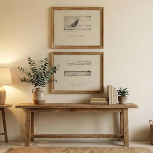 May include: Two framed nautical prints featuring sailboats are displayed above a wooden console table. The table is adorned with a vase of greenery, books, and a potted plant. A jute rug and a lamp complete the scene.