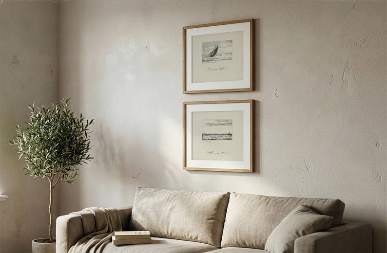 May include: Two framed monochrome landscape prints in wooden frames are displayed above a beige sofa. The prints depict sketches of a sailboat and a coastal scene. A potted olive tree is on the left.