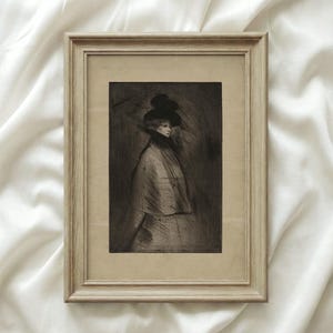 May include: A framed, sepia-toned print of a woman in a large hat and cape. The artwork is in a light-colored wooden frame, set against a white fabric background. The image has a vintage aesthetic.