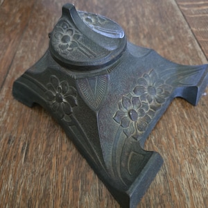 May include: A dark bronze-colored inkwell with floral and leaf designs. The inkwell has a square base with a rounded top and a hinged lid. The lid has a small handle. The inkwell is made of metal and sits on a wooden surface.