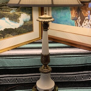 May include: A vintage table lamp with a cream-colored lampshade and a white ceramic base. The lamp features ornate brass accents, including decorative leaves and a square base. The lamp is approximately 30 inches tall.