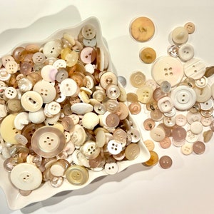 May include: A white ceramic bowl filled with a variety of white and cream colored buttons. The buttons are of different shapes and sizes, and some have a pearl-like finish. Some buttons are scattered on a white surface.