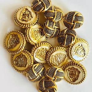May include: A collection of gold-colored buttons. Some buttons have a woven texture, while others feature a cream-colored center with a gold emblem and a decorative gold border. The buttons are arranged on a white surface.
