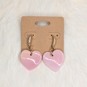 Peut inclure: Une paire de boucles d'oreilles en forme de cœur. Chaque boucle d'oreille présente un pendentif en forme de cœur rose clair suspendu à un anneau doré orné de petites pierres transparentes. Les boucles d'oreilles sont présentées sur une carte marron.