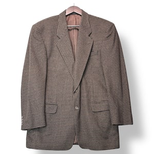 Könnte beinhalten: Braune und schwarze Tweed-Jacke an einem Holzbügel. Die Jacke hat ein fallendes Revers, zwei Vordertaschen und einen Ein-Knopf-Verschluss. Die Ärmel haben drei Knöpfe an der Manschette.
